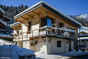 Wooden alpine chalet with a snow-covered roof and balcony in a sunny winter landscape.