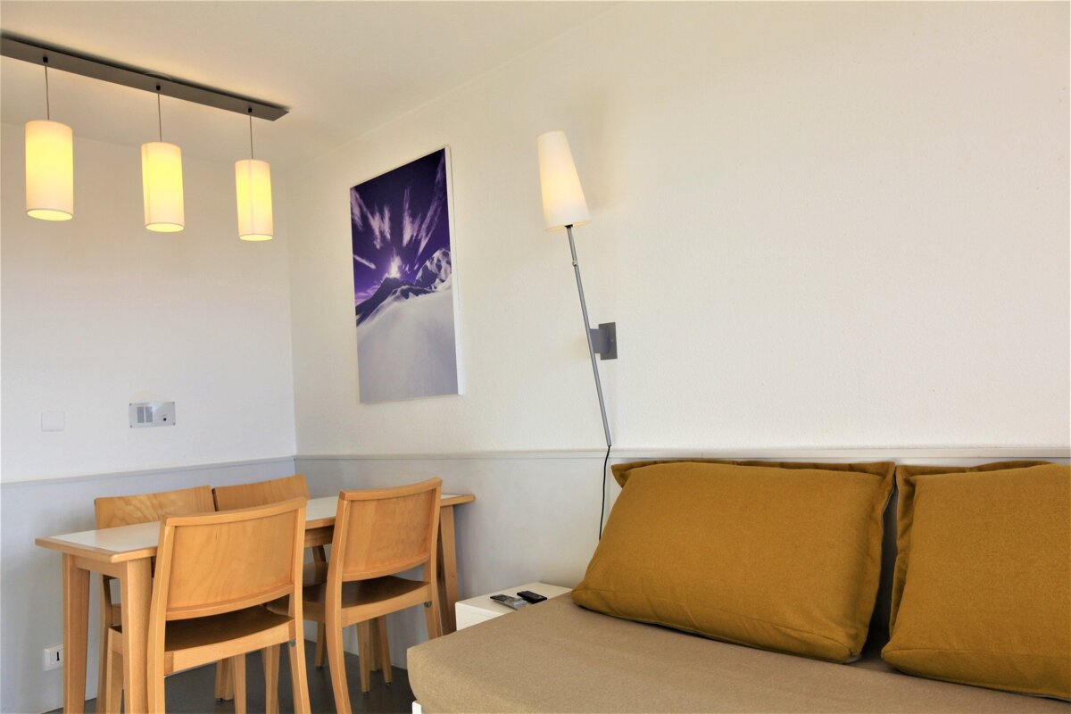 A modern room with a dining table, wooden chairs, a sofa with mustard yellow pillows, and a purple mountain art print on white walls.