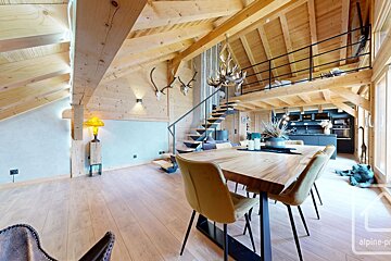 Spacious, modern chalet interior with exposed wood, high ceilings, a dining area, dark kitchen, and loft with a black railing. Antlers decorate walls.