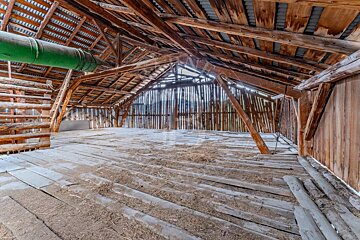 Rustic wooden barn attic with exposed beams, a dirty floor, a green pipe, and sunlight filtering through the wall slats.
