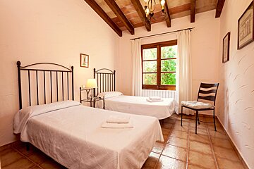 A rustic twin bedroom with wrought iron beds, exposed wooden beams, terracotta floor, and a window offering a view of lush greenery.