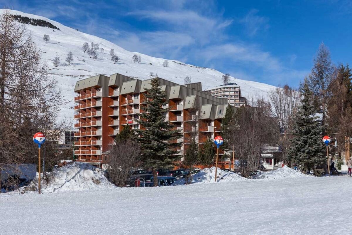 A snowy ski resort with large apartment buildings, pine trees, and distant snow-covered mountains under a bright blue sky.