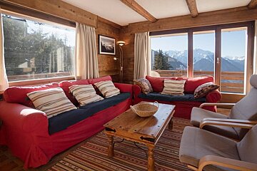 A rustic wooden cabin living room with two red sofas, a wooden coffee table, and a scenic mountain view through large windows.