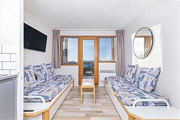 A cozy studio room with two blue-patterned daybeds, a central stool, and large windows revealing a mountain view. A TV and mirror adorn the walls.