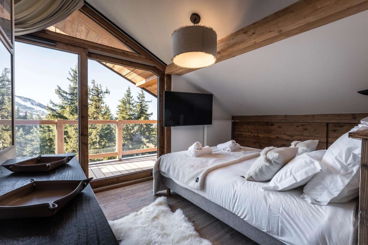 Cozy chalet bedroom with large window and balcony overlooking a mountain and forest. Features a comfy bed, TV, and wooden decor.