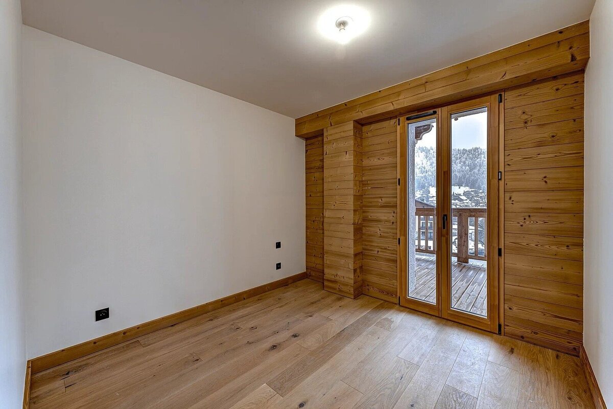 Empty room with wood floors, white walls, and wood-paneled glass doors opening to a balcony with a snowy mountain view.