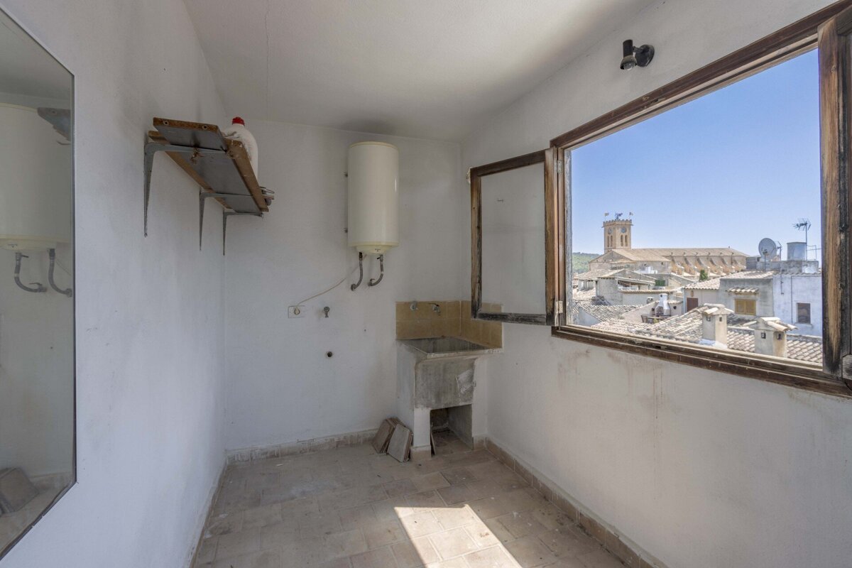 An empty utility room with a sink, water heater, and an open window revealing an old town's rooftops and a prominent church tower under a blue sky.