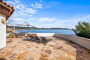 A stone terrace with lounge chairs overlooks a calm blue sea under a bright, partly cloudy sky.