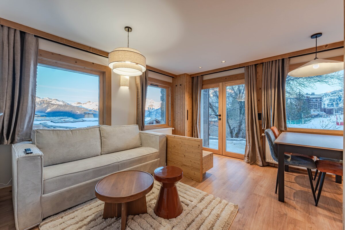 Cozy alpine living room with a beige sofa, dining set, and large windows showing scenic snowy mountain views.