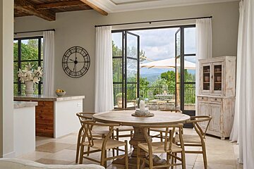 A rustic dining room with a wooden table opens to a scenic balcony overlooking mountains and greenery, featuring a large clock and cabinet.