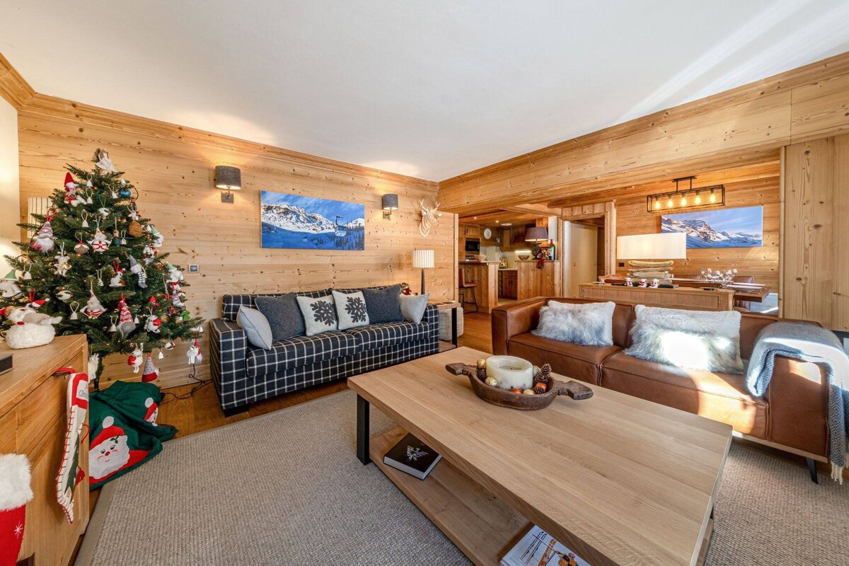A cozy, wood-paneled living room with a decorated Christmas tree, two sofas, a coffee table, and mountain artwork, creating a festive, rustic vibe.