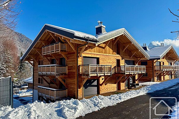 Two wooden chalets with balconies stand in a snowy mountain landscape under a clear blue sky, offering a winter retreat.