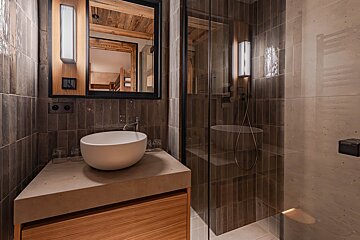 Sleek, modern bathroom with a white bowl sink on a concrete vanity, wood accents, dark tiled walls, and a glass shower. Bunk beds reflect in the mirror.