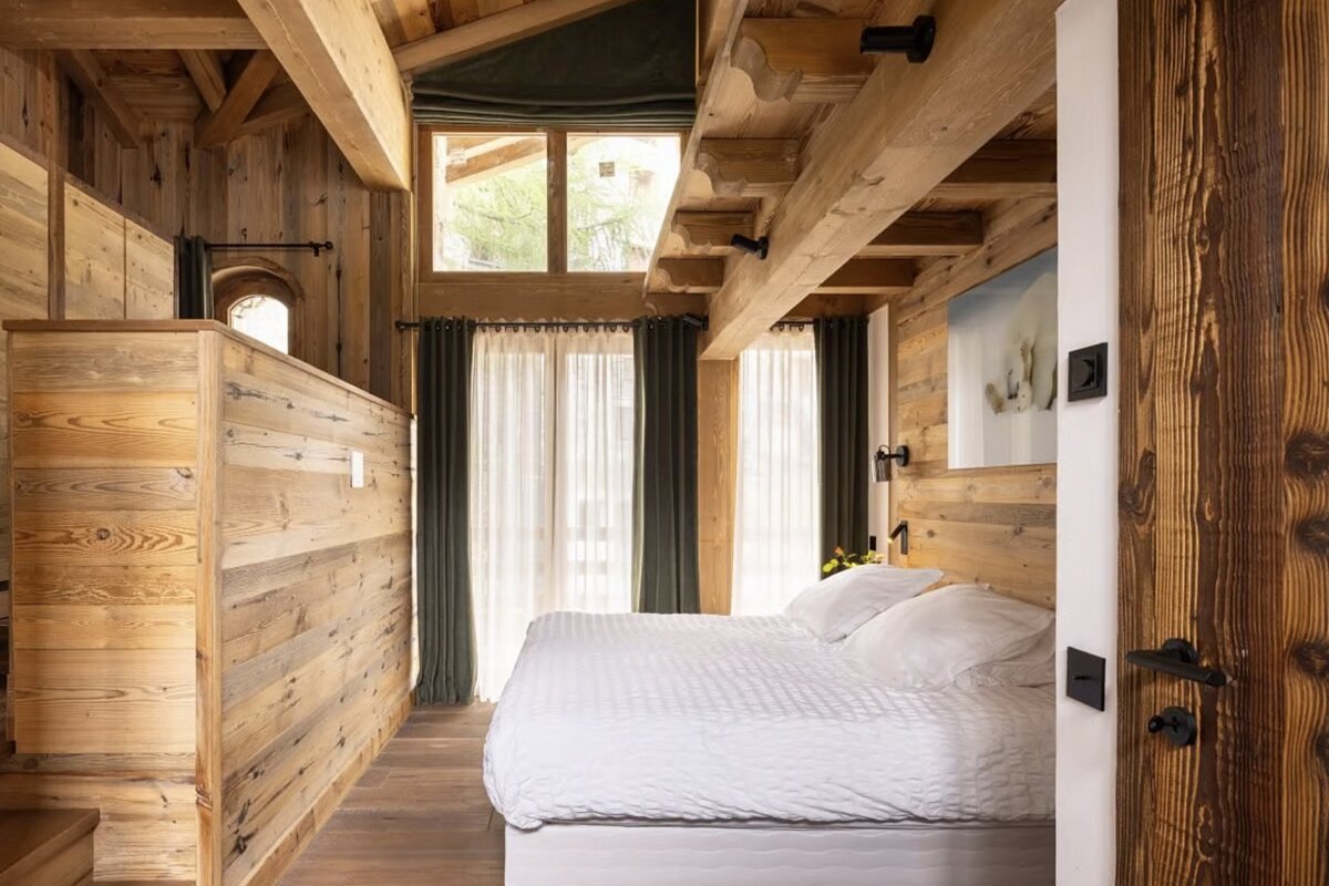A cozy, rustic bedroom featuring warm wooden walls, exposed beams, a white bed, and large windows with sheer and dark curtains.