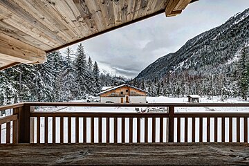 A rustic wooden balcony frames a snowy mountain valley with pine trees, a cozy chalet, and a distant mountain under an overcast sky.