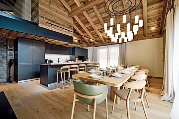 A modern kitchen and dining room with a high, rustic wooden ceiling. Dark cabinetry contrasts with light wood floors and a long dining table.