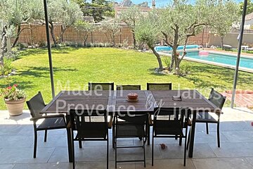 An outdoor patio with a dining table and chairs, overlooking a green lawn, several olive trees, and a swimming pool in the background.