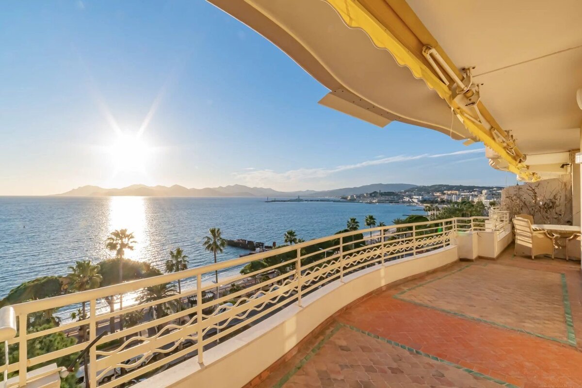 A bright, sunny balcony offers panoramic views of the ocean, a distant coastline, and palm trees below.