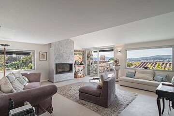 A spacious, bright living room featuring a modern fireplace, comfortable seating, and large windows with panoramic views of a city, hills, and a terrace.