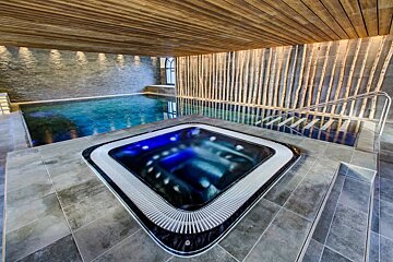 A luxurious indoor spa featuring a glowing blue jacuzzi, a swimming pool, and warm wood and stone finishes.