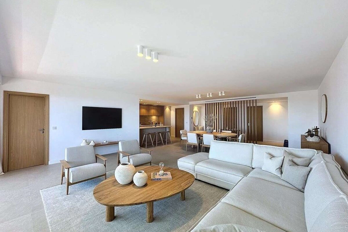 A modern, open-plan living room with a white sectional sofa, wooden coffee table, armchairs, wall-mounted TV, and a kitchen/dining area.