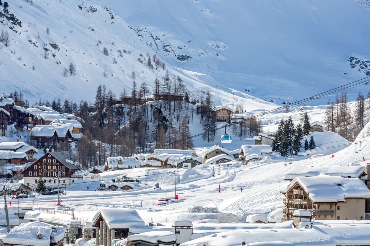 A sunny winter day in a snow-covered alpine village nestled in mountains, featuring chalets, bare trees, ski slopes, and a gondola.