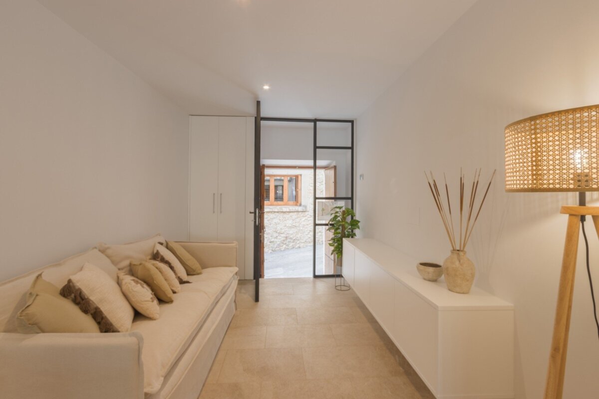 A bright, minimalist room features a beige sofa, white storage, and a black-framed glass door leading to a rustic stone facade.