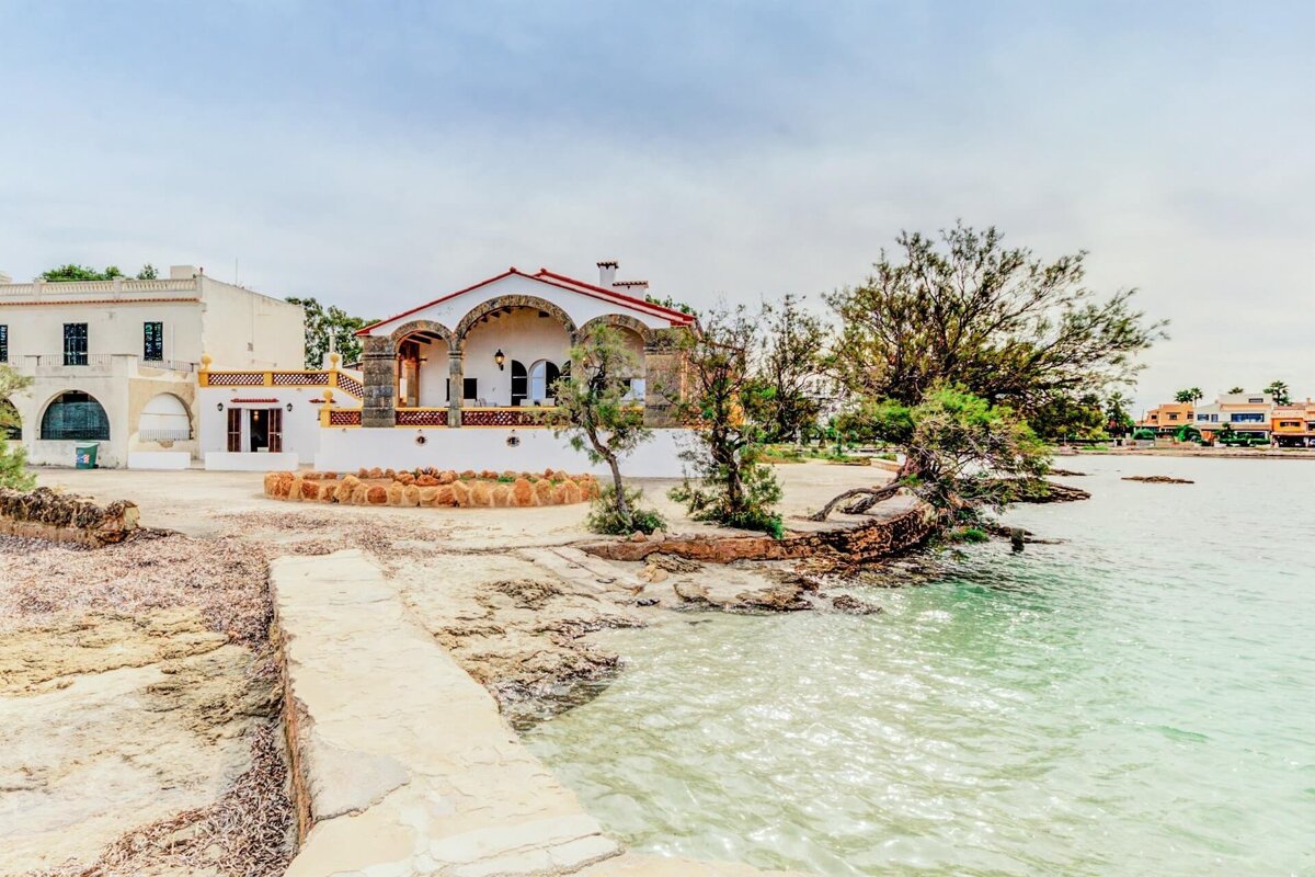 A white house with a red roof and arches sits by clear, turquoise waters. Rocky shoreline, twisted trees, and distant buildings create a peaceful coastal view.