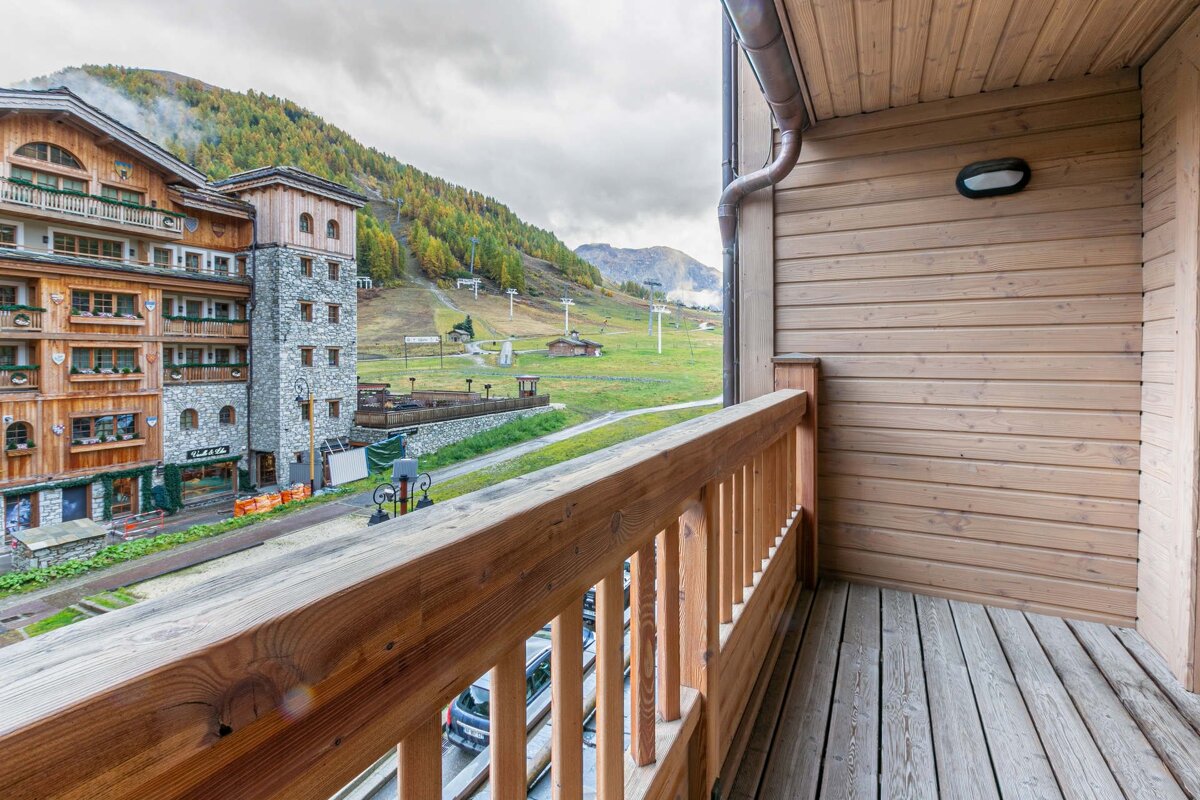 A wooden balcony overlooks a mountain village with rustic buildings, green ski slopes, and lifts under a cloudy sky.