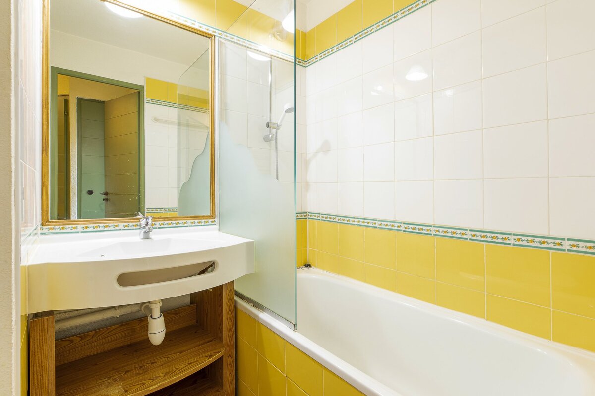A bright bathroom with yellow and white tiled walls, featuring a white sink under a mirror, and a bathtub with a partial glass shower screen.