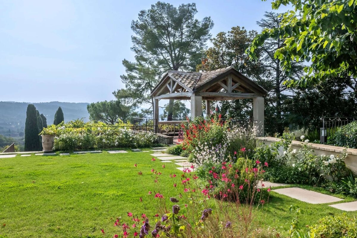 A lush garden features vibrant flowers, a stone path, and a rustic gazebo, overlooking distant hills under a clear sky.