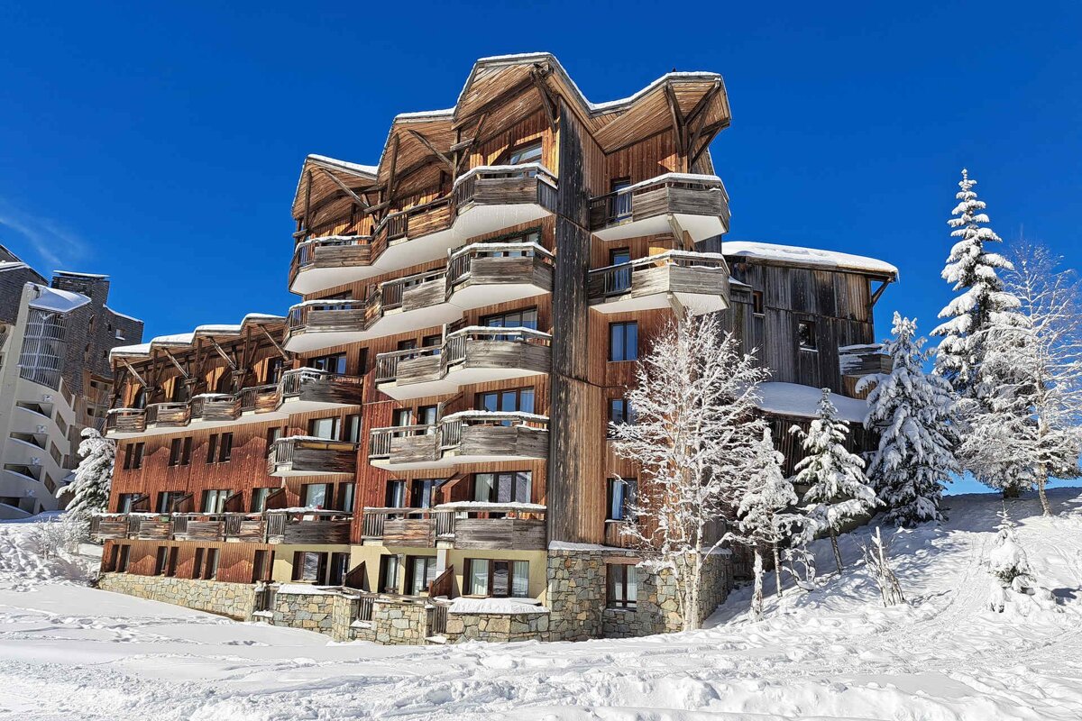 A snow-covered multi-story wooden chalet with many balconies and surrounding snowy trees, under a bright blue sky.