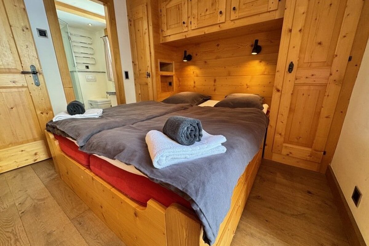 Cozy wooden chalet bedroom featuring a double bed with grey covers, folded towels, built-in storage, and a view of an adjacent bathroom.