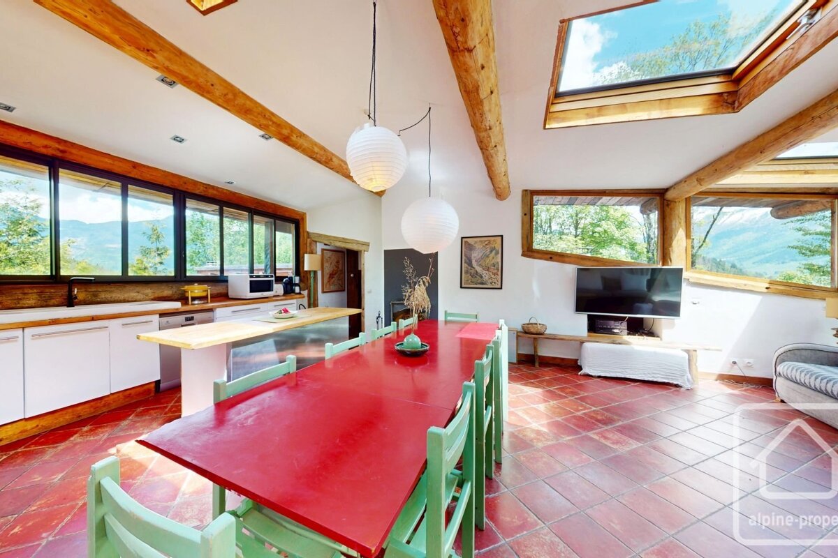 A bright chalet interior with a long red dining table, terracotta tile floor, wooden beams, skylight, and large windows.