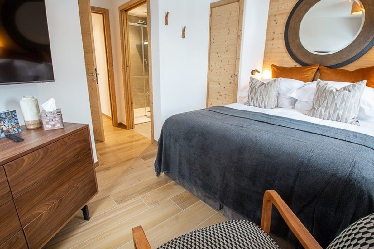 A cozy modern bedroom featuring a bed with a grey throw, a large round mirror, wooden dresser, and an open doorway to a bathroom.