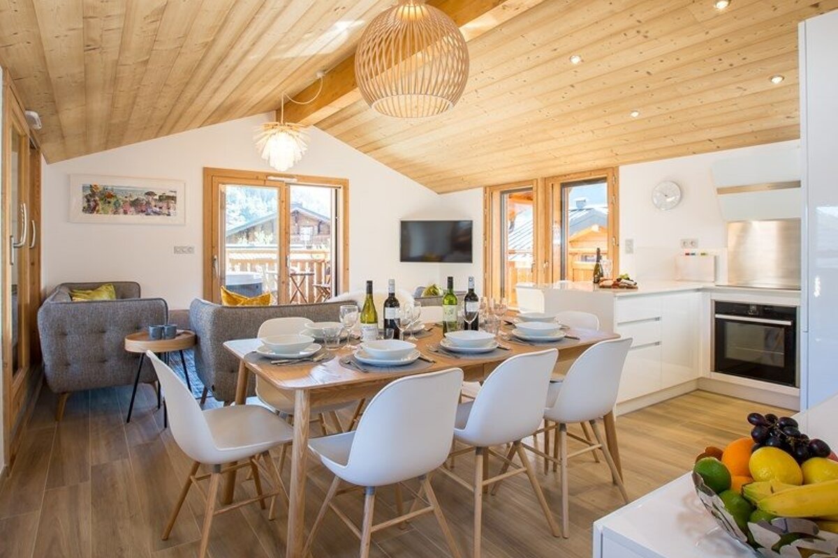 Bright, open-plan chalet interior with a wooden dining table, white chairs, kitchen, and living area under a sloped wood ceiling.