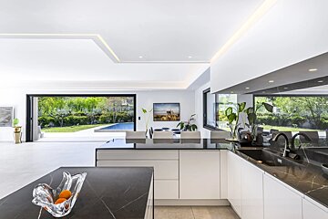 A modern, white kitchen featuring black stone countertops, with glass doors opening to a backyard pool.