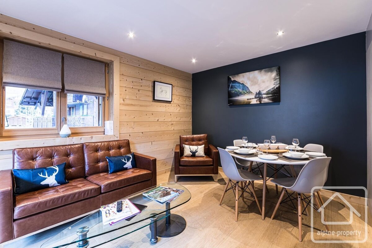 Modern chalet interior with a brown leather sofa, glass coffee table, and a dining area set against a dark accent wall.