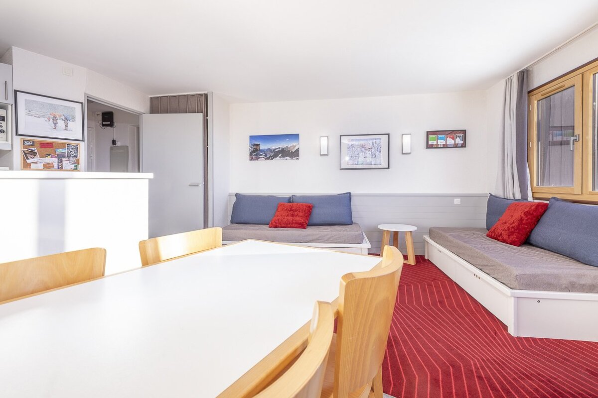 Bright living/dining room with a red geometric carpet, white dining table, and wooden chairs. Two daybeds with blue cushions and red pillows are against white walls adorned with mountain art.
