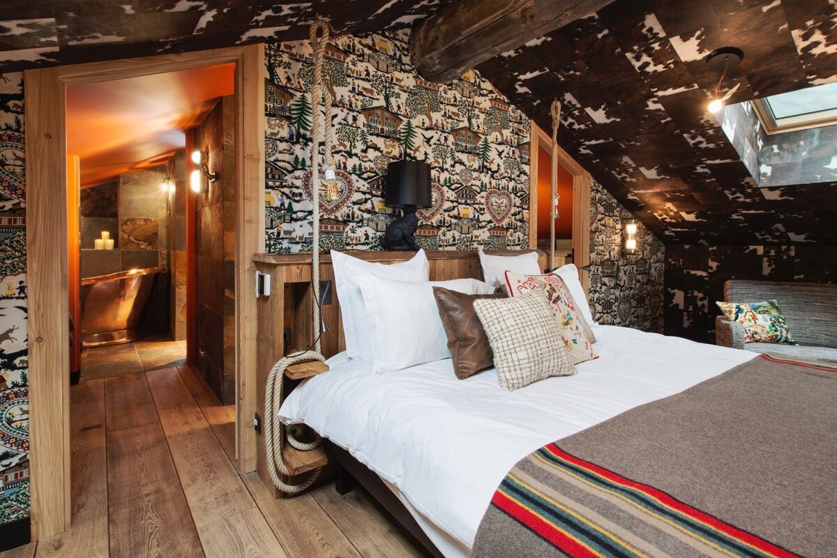 A rustic bedroom with patterned alpine wallpaper, rope-detailed bed, wood floors, cowhide ceiling, and an ensuite bathroom with a copper tub.