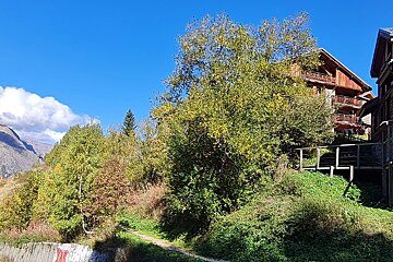 Wooden chalets on a lush, tree-covered hillside under a clear blue sky, with a mountain peak visible in the distance.