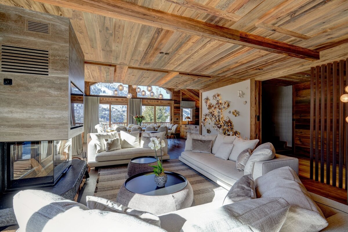 A luxurious, modern living room with warm wooden ceilings, light furniture, a sleek fireplace, and large windows showcasing a stunning snowy mountain view.