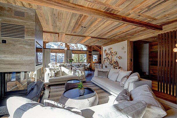 A luxurious, modern living room with warm wooden ceilings, light furniture, a sleek fireplace, and large windows showcasing a stunning snowy mountain view.