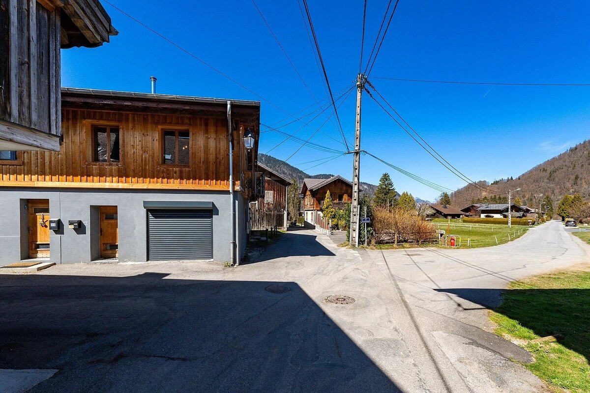 Sunny village street with wooden buildings, a utility pole, and a winding road leading to green hills under a clear blue sky.