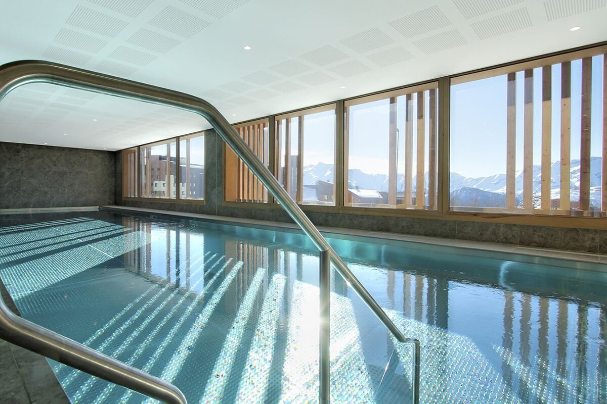 A bright indoor swimming pool with large windows overlooking snow-capped mountains. Sunlight creates shimmering patterns on the blue water.