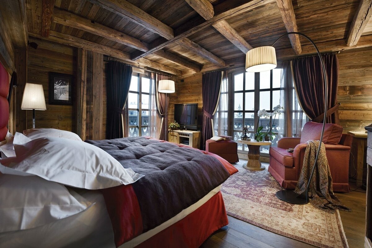 Luxurious rustic bedroom with a large bed, wooden walls, beamed ceiling, armchairs, and expansive windows offering a view.