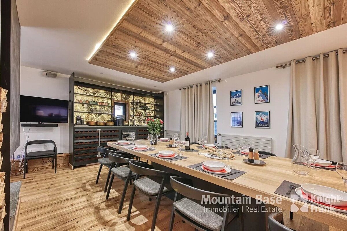 A chic dining room with a long wooden table set for guests, featuring a stylish bar with wine racks and glasses, a wooden ceiling with spotlights, and ski posters on the wall.
