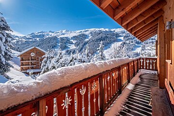 View from a snowy wooden balcony over a winter wonderland: charming chalet, snow-covered pine trees, and majestic mountains under a blue sky.