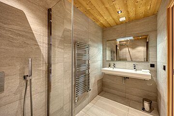 Modern bathroom featuring light grey tiles, a sloped wooden ceiling, glass shower, towel warmer, and a double vanity with mirror.