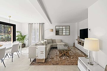 A bright, open-plan living and dining room with a light L-shaped sofa, rustic coffee table, dining set, and large windows.